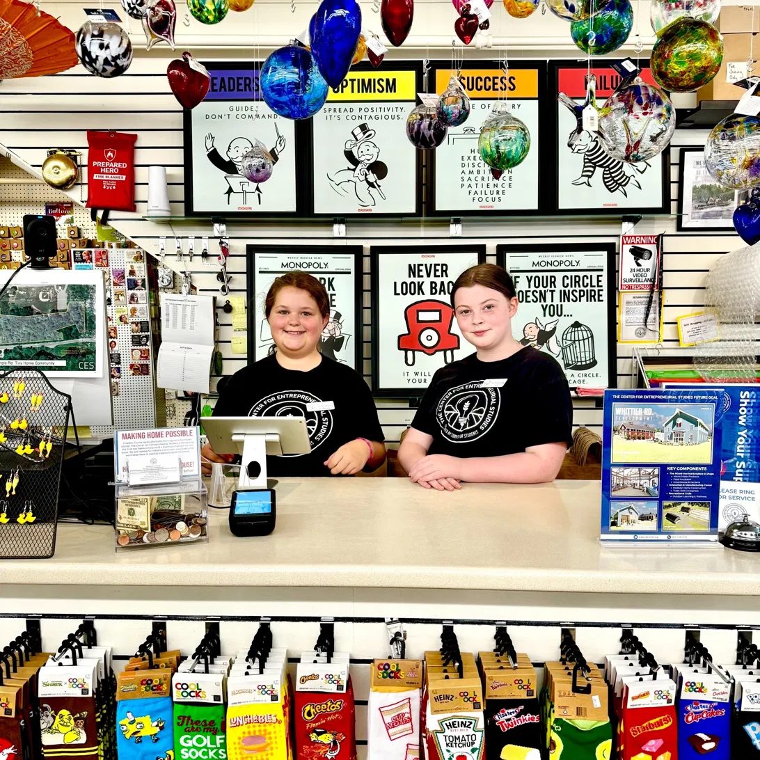 Two young people in CES T-shirts behind the counter of the CES store on Main Street, hand-blown glass ornaments hanging above them.